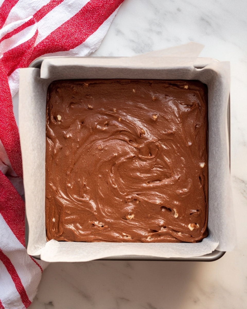 The image shows a square metal baking pan lined with parchment paper filled with a smooth, thick layer of chocolate batter that has small nut pieces mixed throughout. The batter is evenly spread inside the pan with slight swirls on the surface, giving it a textured look. The pan is placed on a white marbled surface with a folded red and white kitchen towel visible on the left side. Photo taken with an iphone --ar 4:5 --v 7