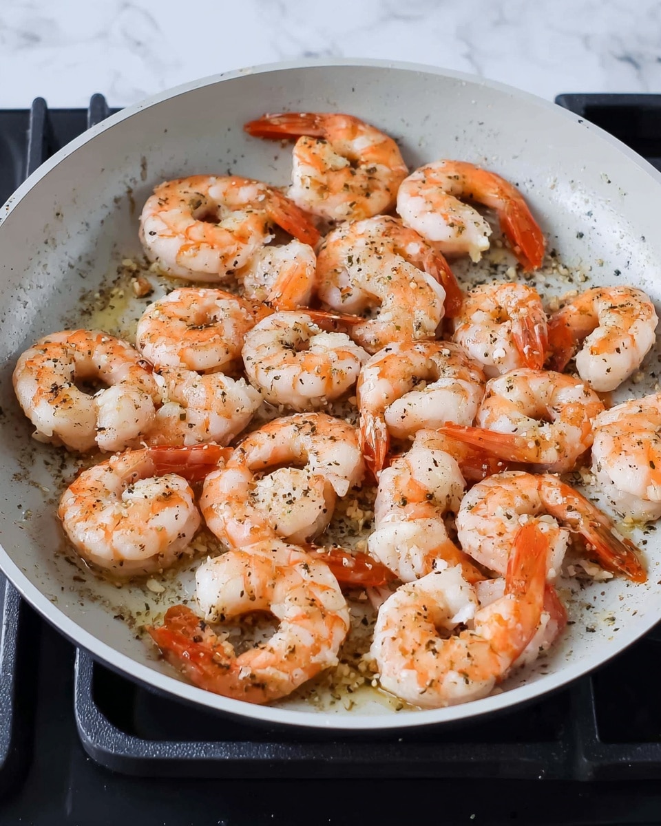 A black pan filled with creamy pasta showing multiple layers: long, thick spaghetti noodles twisted through a rich white sauce that looks smooth and thick, dotted with many medium-sized shrimp that are pale pink with slight orange touches. Scattered green herbs and small bits of what looks like sun-dried tomatoes add color contrast throughout. The sauce appears slightly speckled with black pepper, giving texture. The pan sits on a white marbled surface with a stove edge slightly visible in the background photo taken with an iphone --ar 4:5 --v 7