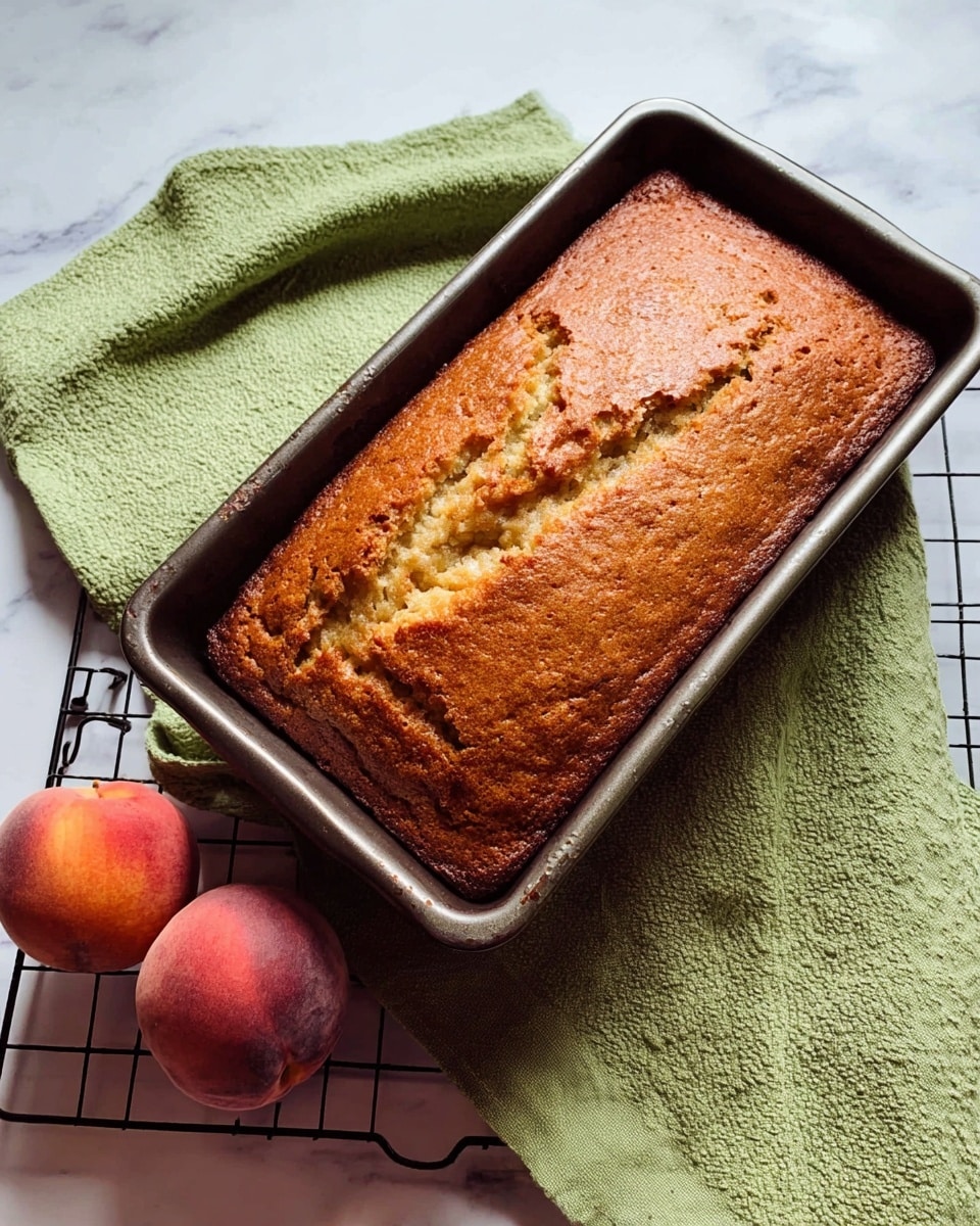 A golden-brown loaf cake sits in a dark metal rectangular baking pan with a slightly cracked top surface showing its soft, textured inside. The pan rests on a green textured cloth, placed over a white marbled surface. Beside the pan, two whole ripe peaches with a fuzzy skin in shades of red and yellow lie on a black wire cooling rack, adding a fresh fruit contrast to the baked cake. photo taken with an iphone --ar 4:5 --v 7
