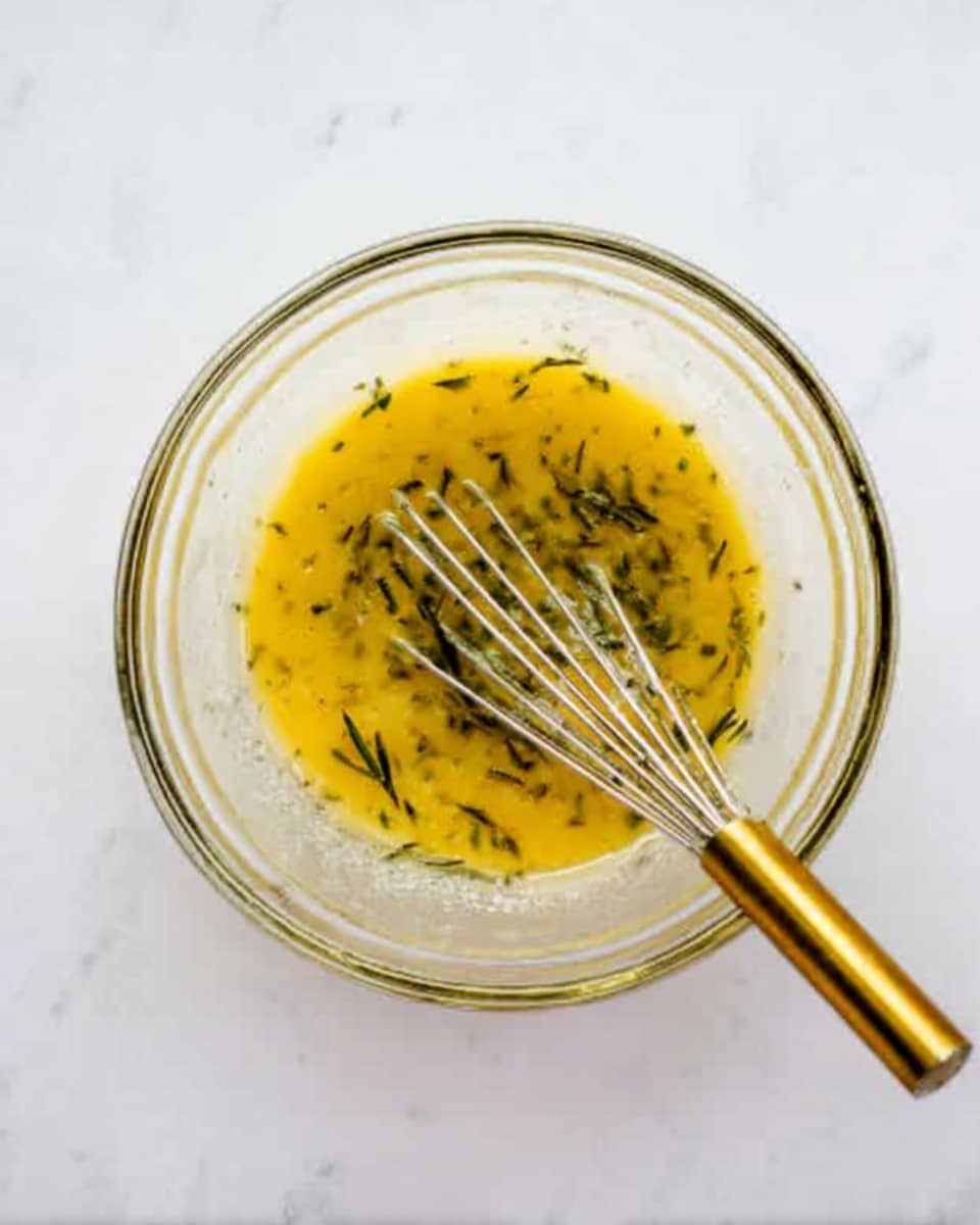 A clear glass bowl filled with a yellow sauce that has small green herb pieces mixed throughout. Inside the bowl, a whisk with a gold handle is partially dipped into the sauce. The bowl is placed on a white marbled surface. photo taken with an iphone --ar 4:5 --v 7