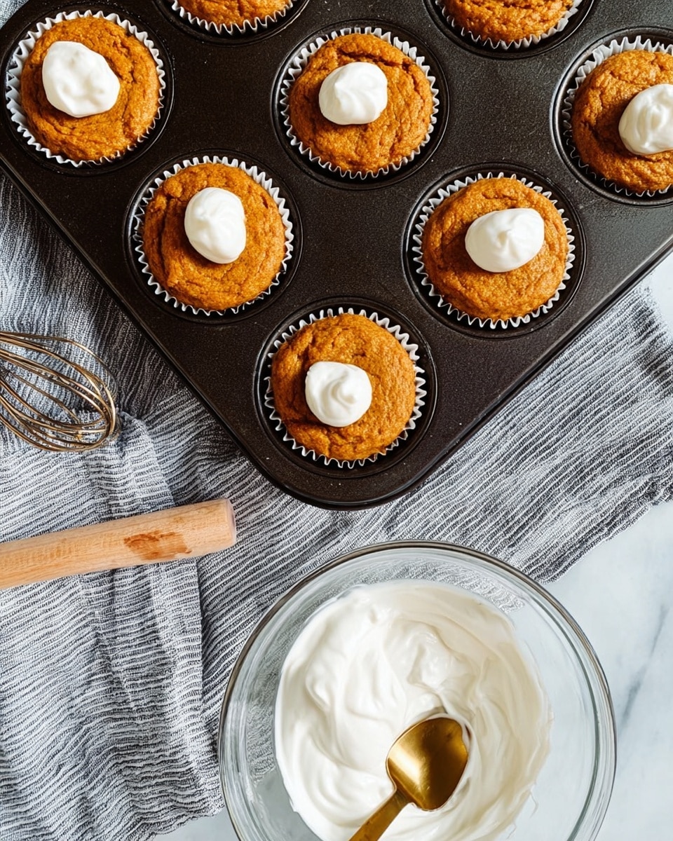 The image shows two golden brown muffins stacked on top of each other, each wrapped in white paper liners with a dollop of creamy white frosting on top. In the foreground, there is a partially eaten muffin revealing its soft, moist inside with some frosting on the exposed part. To the right, a silver knife lies flat on a light surface. In the background, additional muffins sit in a black muffin tray and on the table, with a white marbled surface underneath. A blurred striped cloth and a small decorative pumpkin add depth to the scene. Photo taken with an iphone --ar 4:5 --v 7