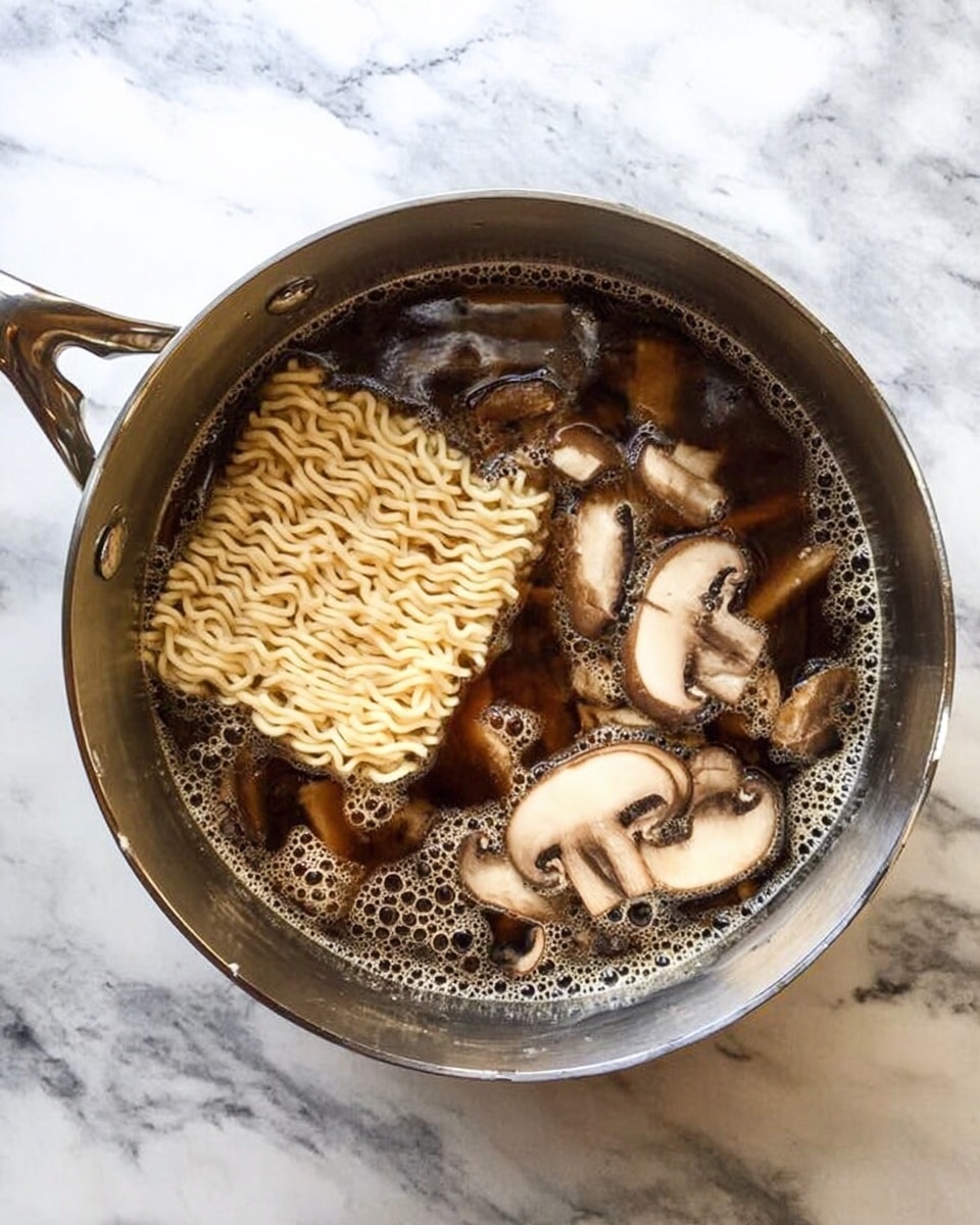 A top view of a silver pot on a white marbled surface filled with dark brown broth, sliced light brown mushrooms floating on the right side, and a block of uncooked pale yellow instant noodles partially submerged on the left side, with bubbles forming around the edges of the soup photo taken with an iphone --ar 4:5 --v 7