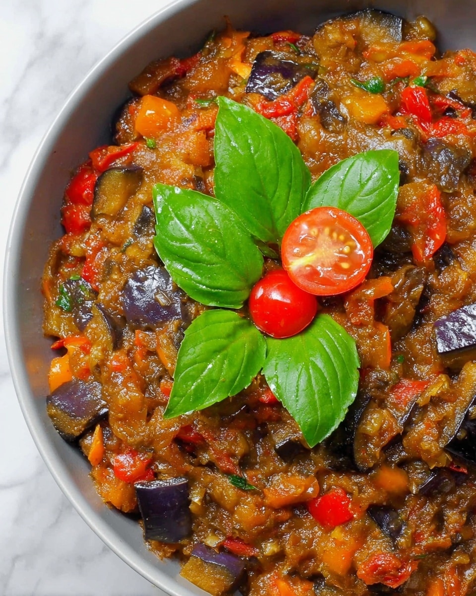 The dish is a mixed vegetable stew with a soft, chunky texture combining brown, orange, and purple colors from cooked eggplants, red and orange bell peppers, and onions. On top, there are five fresh, bright green basil leaves arranged around a small red cherry tomato sliced in half, showing its seeds. The stew fills a white bowl with a close-up view, and the background is a white marbled texture. photo taken with an iphone --ar 4:5 --v 7