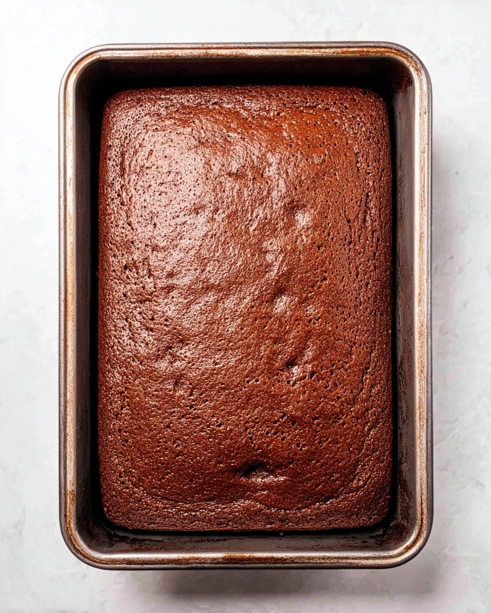 A clear glass bowl shows a layered dessert with three main layers. The bottom layer is a crumbly, dark brown layer. The middle layer is a thick, smooth, light cream-colored layer. On top of that, there is a layer of dark brown cake cubes. Above the cubes, a thick, pale cream layer is being spread with a white spatula held by a woman's hand. The background has a white marbled texture with small red berries blurred in the lower part of the image. photo taken with an iphone --ar 4:5 --v 7