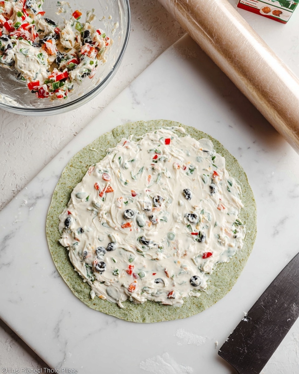 A flat green spinach wrap lies on a white marbled surface, spread evenly with a creamy white mixture that has visible small pieces of black and green olives and red bell pepper bits scattered throughout. Above the wrap, a clear glass bowl contains more of the creamy mixture with mixed olives and peppers. To the right, a roll of cling film in a box sits on the white marbled surface, and below it, a knife with a black handle rests near the wrap. The scene is bright and clean, showing the fresh ingredients and preparation in progress. photo taken with an iphone --ar 4:5 --v 7