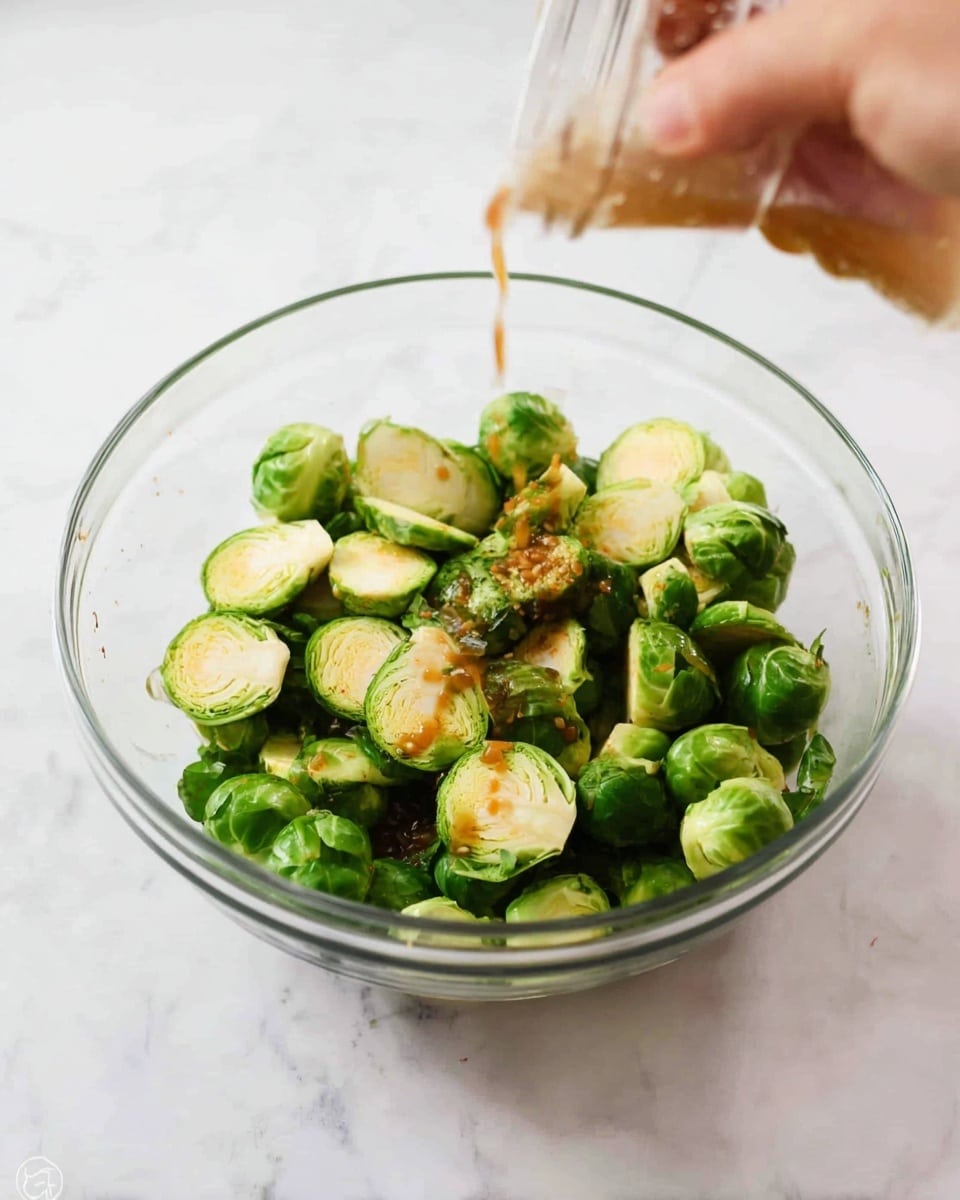 A clear glass bowl filled with two layers of halved small green Brussels sprouts showing a mix of light and darker green leaves. A woman's hand is pouring golden-brown sauce over the Brussels sprouts, creating a shiny wet texture on some pieces. The bowl sits on a white marbled surface, and the image is bright and clean with soft shadows. photo taken with an iphone --ar 4:5 --v 7