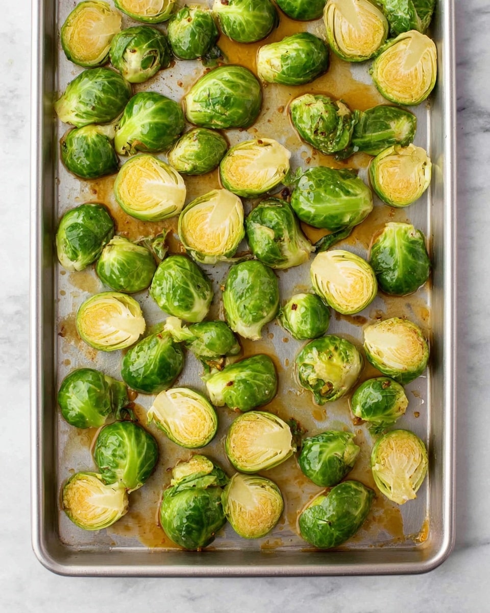 The image shows a metal baking tray filled with halved Brussels sprouts scattered evenly across the tray. Each Brussels sprout is bright green with light yellow inside, some showing slight browning from roasting. There is a layer of golden brown sauce or oil pooled in spots on the tray and around the sprouts, adding a slight shine to the vegetables. The background is a white marbled surface, giving a clean and simple look to the scene. photo taken with an iphone --ar 4:5 --v 7