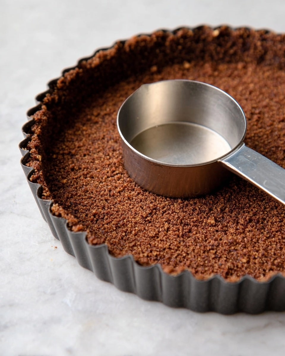 A close-up image of a brown crumb crust pressed firmly inside a dark metal tart pan with fluted edges, showing the textured crumb layer evenly spread with a slightly raised edge all around; a shiny silver measuring cup lies inside the tart pan on top of the crust, contrasting with the coarse crumb surface beneath it, all placed on a white marbled background photo taken with an iphone --ar 4:5 --v 7