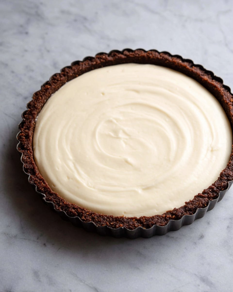 A tart is shown with two layers on a white marbled surface: the bottom layer is a thick, rough-textured dark brown crust forming the edge and base of the tart, while the top layer is a smooth, creamy off-white filling spread evenly to the tart’s edges, with gentle swirls visible on the surface. The tart is in a dark metal tart pan with scalloped edges. Photo taken with an iphone --ar 4:5 --v 7
