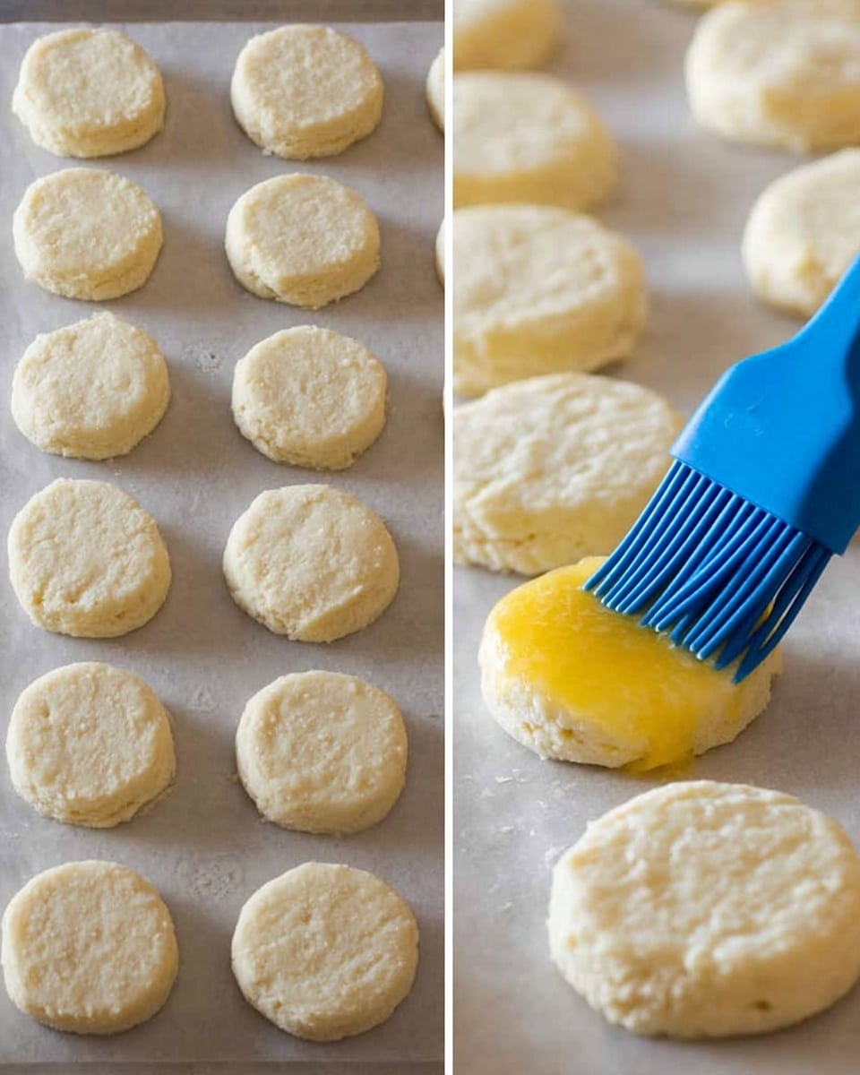 The image shows two side-by-side close-up views of raw biscuit dough pieces arranged neatly on a light-colored baking sheet. Each biscuit is round, pale, and soft with a slightly uneven surface texture from being gently pressed. On the right side, a blue silicone brush is shown spreading a shiny yellow layer of butter or egg wash on one of the biscuits. The biscuits are placed evenly, with soft shadows between them, all set on a white marbled background. photo taken with an iphone --ar 4:5 --v 7