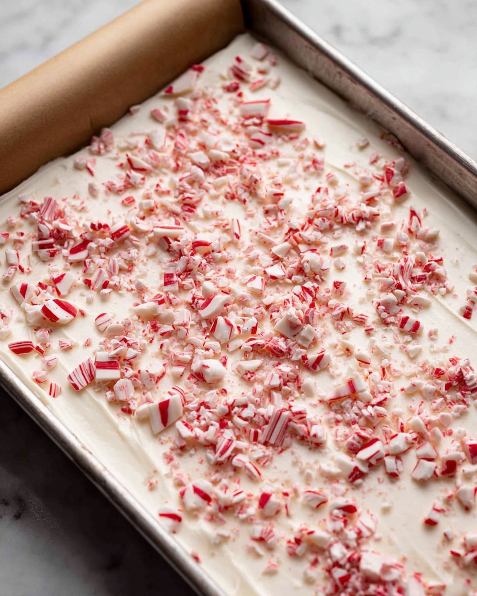 A metal tray holds a thick, smooth white layer resembling soft cream or yogurt spread evenly across the surface. This white layer is topped with small, broken pieces of red and white peppermint candy scattered generously, adding a rough texture and vibrant color contrast. The tray is partially covered at the top by rolled parchment paper, and the background shows a subtle white marbled texture. photo taken with an iphone --ar 4:5 --v 7