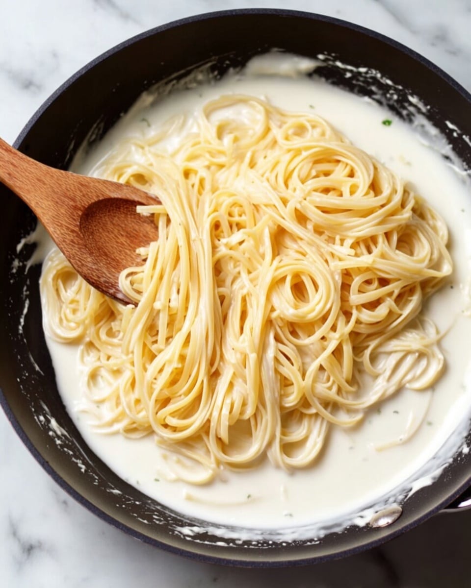 A mound of creamy pasta sits in the center of a white plate with a light blue rim, layered with long, thin noodles coated in a smooth, white sauce. The pasta is topped with finely grated cheese that adds a soft, powdery texture and sprinkled with small green herb pieces that bring a fresh note. The sauce pools slightly around the edges of the pasta, giving a glossy look. The plate rests on a white marbled surface with a blurred background featuring a cheese block and green herbs, making the dish the main focus. photo taken with an iphone --ar 4:5 --v 7