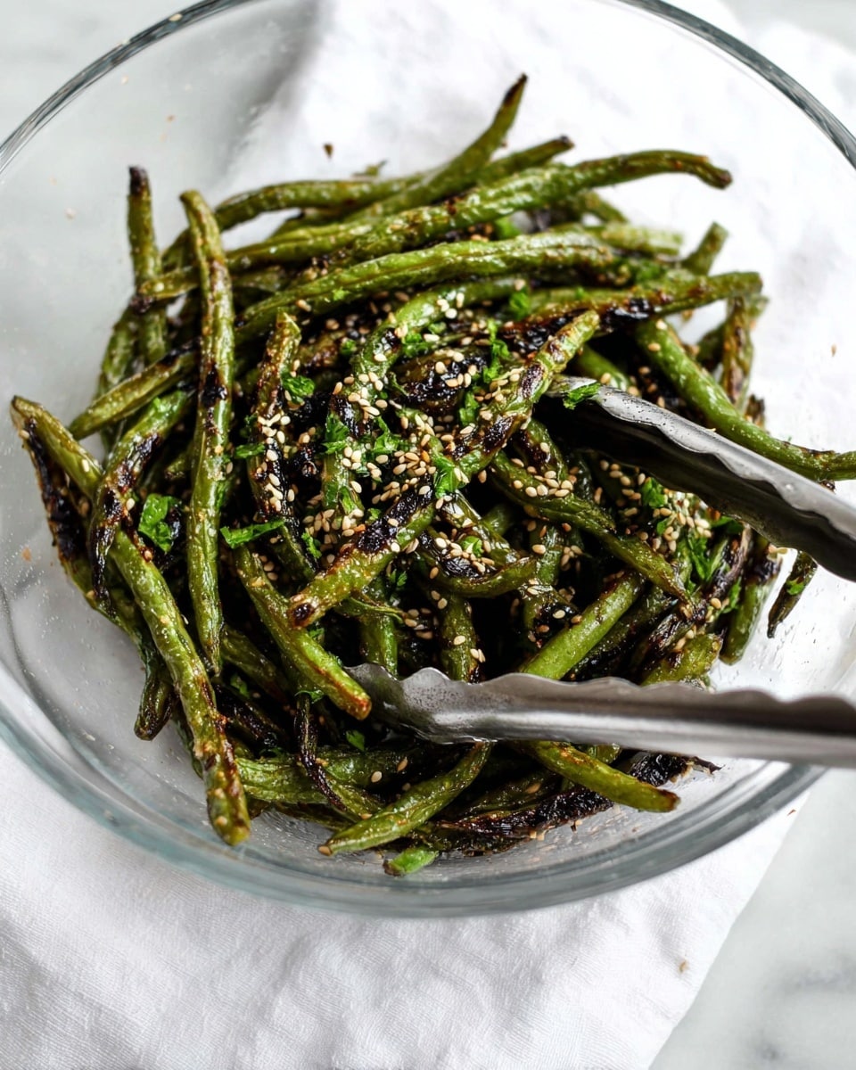 A large white bowl is filled with roasted green beans that have a mix of bright green and dark charred spots showing a crispy texture. The beans are sprinkled with small white sesame seeds and some red chili flakes, adding extra texture and color. Two wooden chopsticks rest on top of the green beans, with one lighter and one darker in wood tone, positioned from the top center going diagonally into the bowl. The bowl sits on a white marbled surface, with a small white wooden dish holding coarse salt at the bottom left and a small round blue bowl filled with green seasoning in the top right background. Bright yellow flowers in a glass vase are partially visible at the top left. photo taken with an iphone --ar 4:5 --v 7