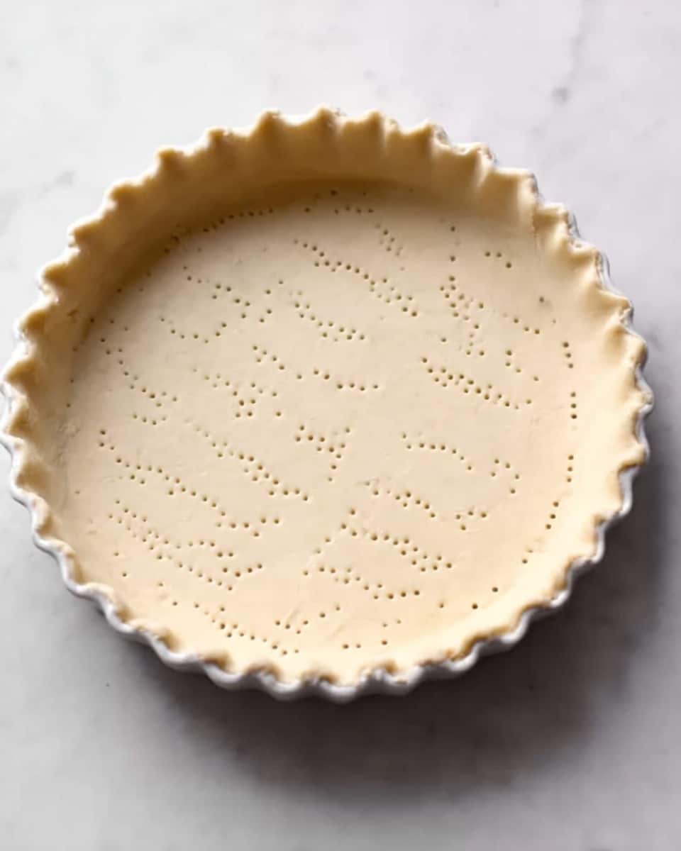 A white pie dish filled with smooth dough that is evenly spread out inside, with small holes poked all over the bottom and sides. The edges of the dough are slightly crimped, forming a wavy pattern around the rim. The dish sits on a white marbled surface. Photo taken with an iphone --ar 4:5 --v 7