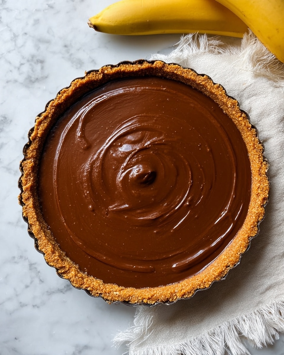 A round tart has a thick, crumbly light brown crust around its edge and holds a smooth, glossy dark brown filling that looks creamy and rich. The surface of the filling shows slight swirls and a small dollop in the center, giving it a soft texture. The tart sits on a white marbled surface next to a yellow banana partly in view at the top right, with a white fringed cloth lying nearby. photo taken with an iphone --ar 4:5 --v 7