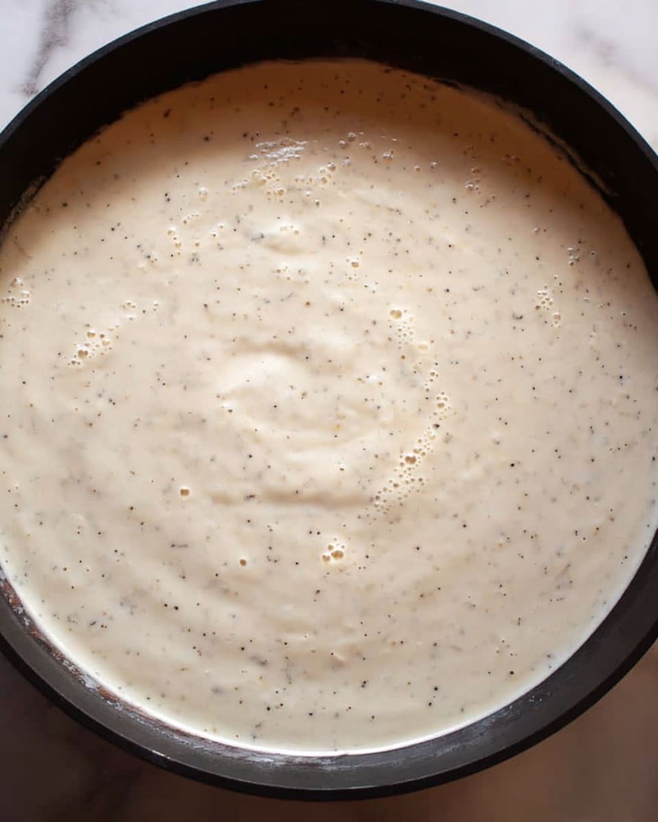 A close-up view of a pan filled with a creamy white sauce that has small black specks scattered throughout, giving it a slightly textured look. The sauce appears thick and smooth with a few small bubbles on the surface, showing it's hot and cooking. The pan has a black edge and is placed on a white marbled surface. Steam gently rises from the sauce, indicating it is warm. Photo taken with an iphone --ar 4:5 --v 7