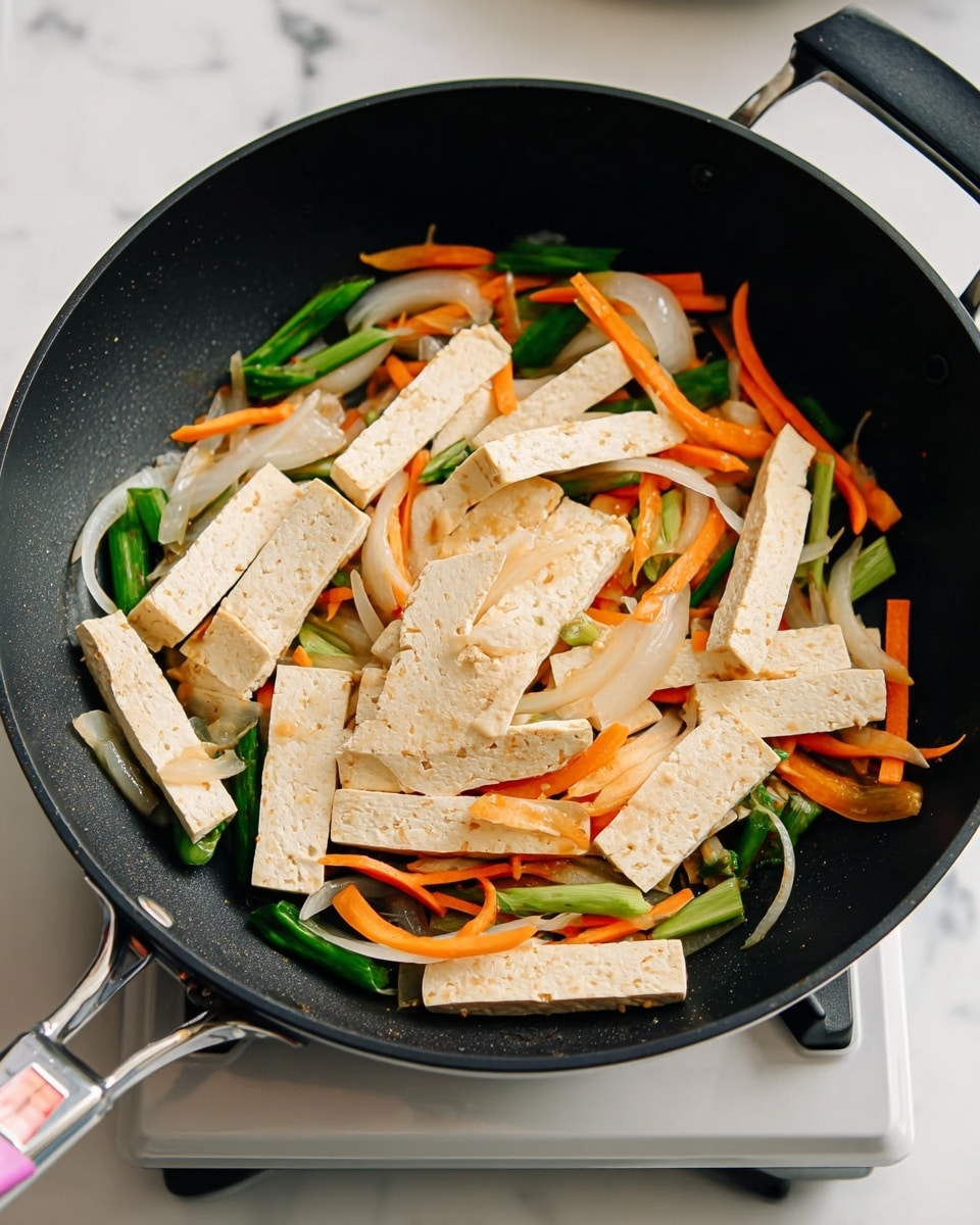 A black frying pan with light beige tofu strips spread across the pan. There are thin white onion slices mixed with thin orange carrot strips and vibrant green spring onion pieces scattered across and under the tofu. The pan is on a small portable stove set on a smooth white marbled surface. The scene is bright and clear, showing the fresh cooked vegetables and tofu in different colors and textures. photo taken with an iphone --ar 4:5 --v 7