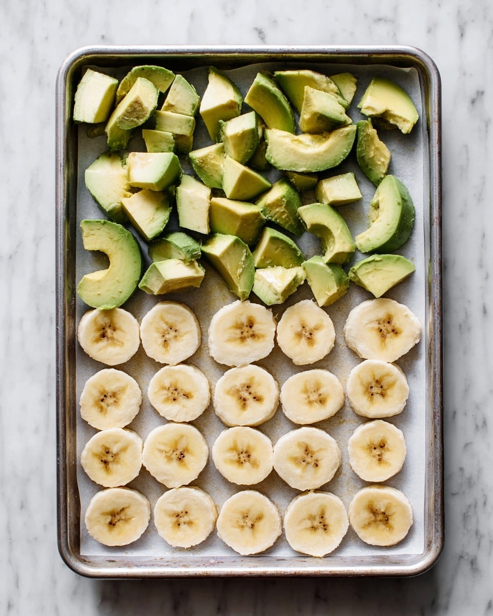 A metal baking tray lined with white parchment paper holds two layers of fruit pieces neatly arranged. The top layer consists of roughly cut avocado chunks, showing a mix of dark green skin and light green flesh with a creamy texture. Below them, there are three rows of round banana slices, each piece pale yellow with a soft texture and small brown specks in the center. The tray is placed on a white marbled surface. photo taken with an iphone --ar 4:5 --v 7