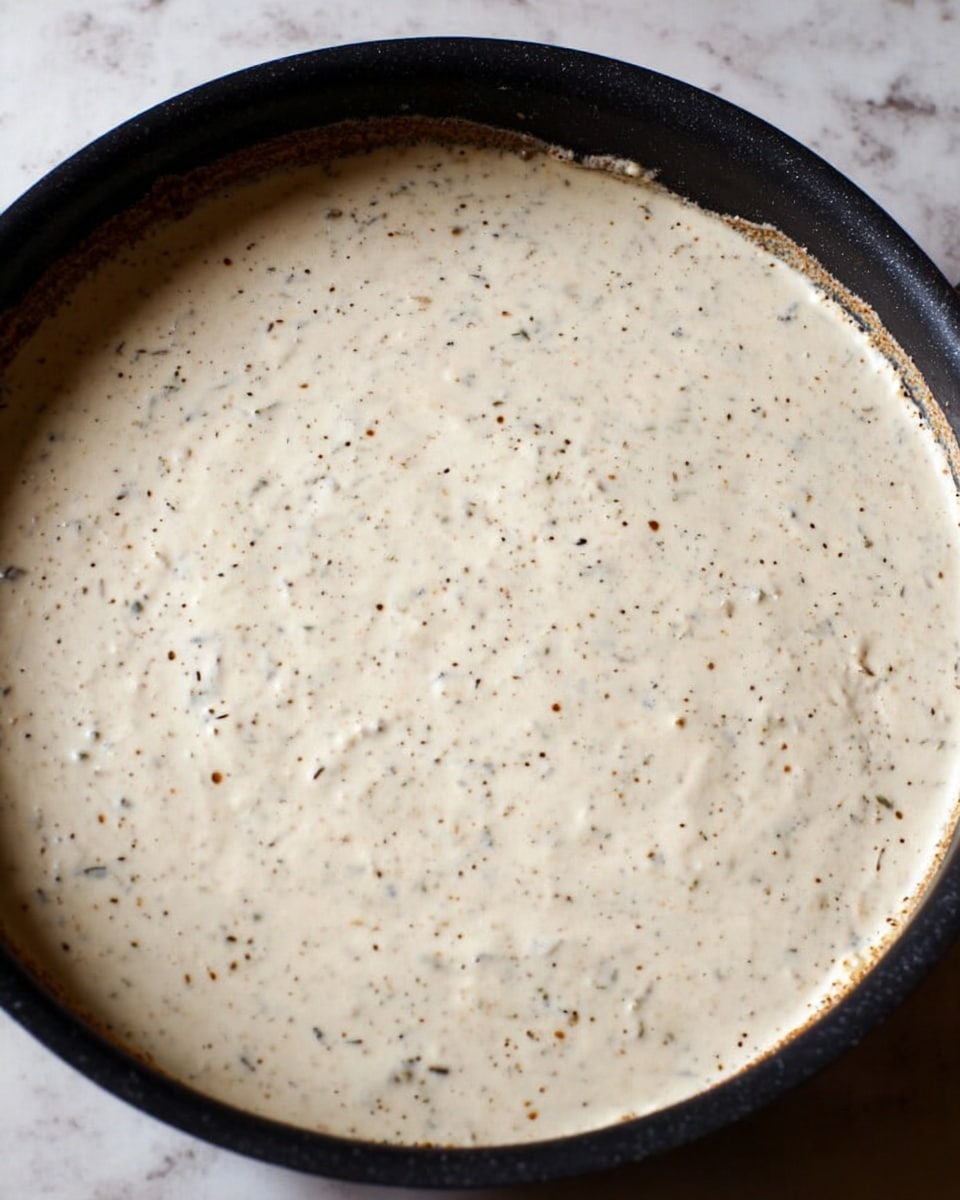 The image shows a close-up of a black pan filled with a creamy white sauce that has specks of herbs and spices scattered evenly throughout. The sauce has a smooth, thick texture and covers the entire visible surface of the pan, with some sauce residue along the pan’s edges. The background is a white marbled surface. photo taken with an iphone --ar 4:5 --v 7
