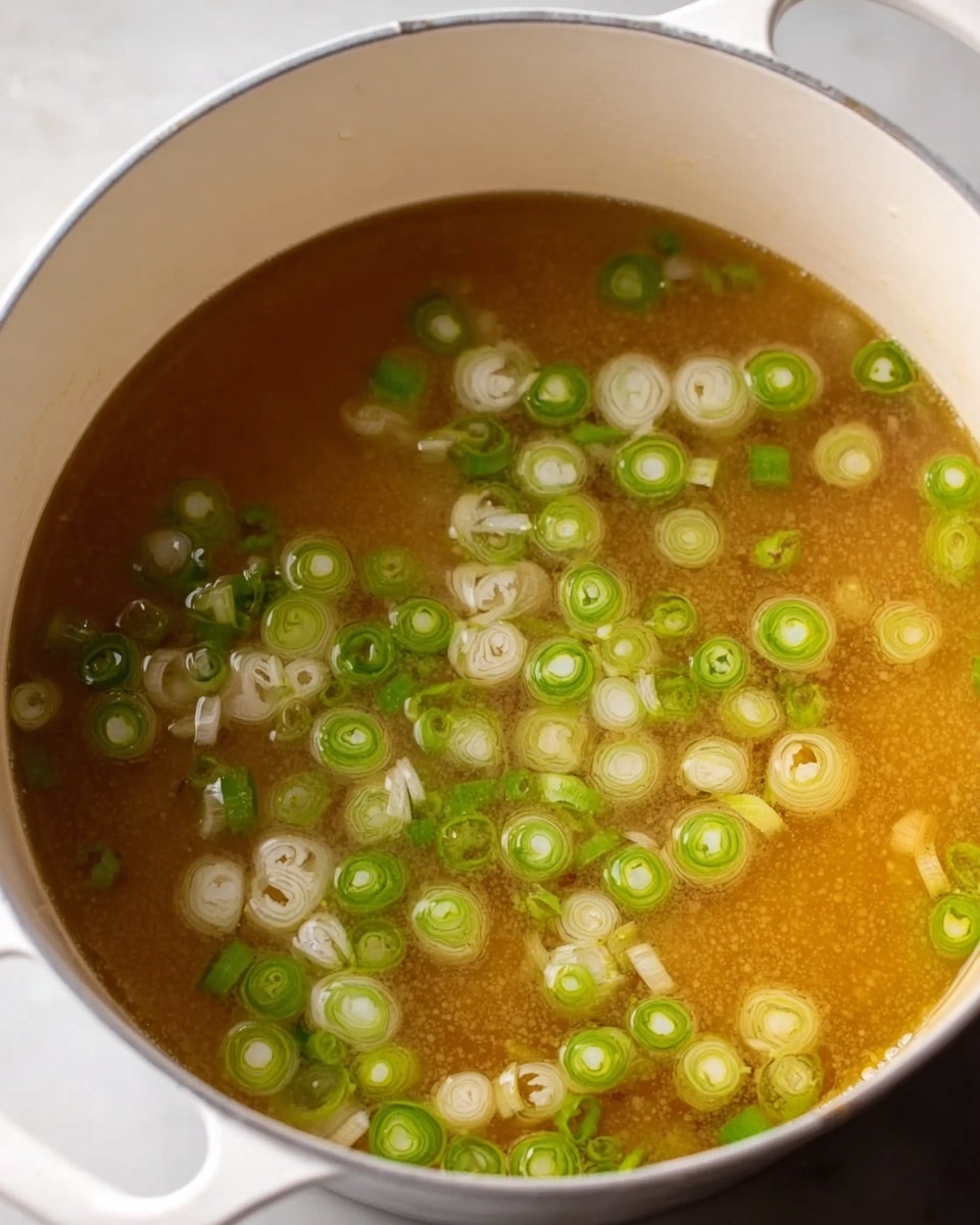 A white pot filled with a light brown broth, showing small bubbles on the surface indicating gentle simmering, and scattered slices of green onions floating on top, creating pops of green and white color against the broth, all placed on a white marbled surface, photo taken with an iphone --ar 4:5 --v 7