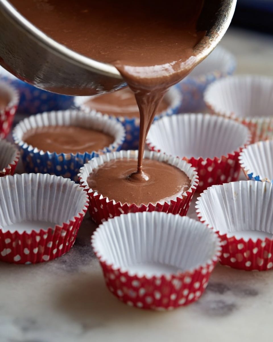 A close-up image shows smooth, light brown chocolate batter being poured from a metal bowl into small, white cupcake liners with red and blue polka dots, arranged in a white marbled tray. The focus is on the glossy liquid flowing steadily into the lined cups, each filled about three-quarters full, highlighting the rich texture of the chocolate batter. In the background, more empty white cupcake liners are visible, creating a sense of preparation. The surface beneath is a white marbled texture, giving a clean and bright contrast to the warm colors of the batter and liners. photo taken with an iphone --ar 4:5 --v 7