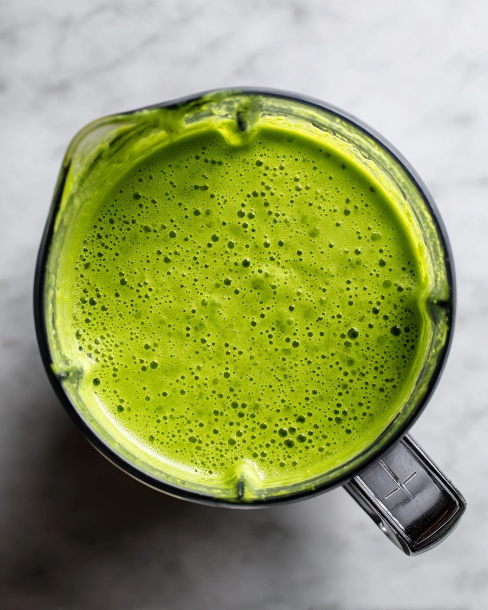 The image shows a clear blender container filled with a thick, bright green smoothie. The smoothie has a smooth texture with small bubbles and tiny darker green specks spread evenly throughout. The blender is placed on a white marbled surface, and the top view captures the wide round opening and the blender's spout shape clearly. The handle of the blender is visible on the right side, highlighting its metallic and black grip design. photo taken with an iphone --ar 4:5 --v 7
