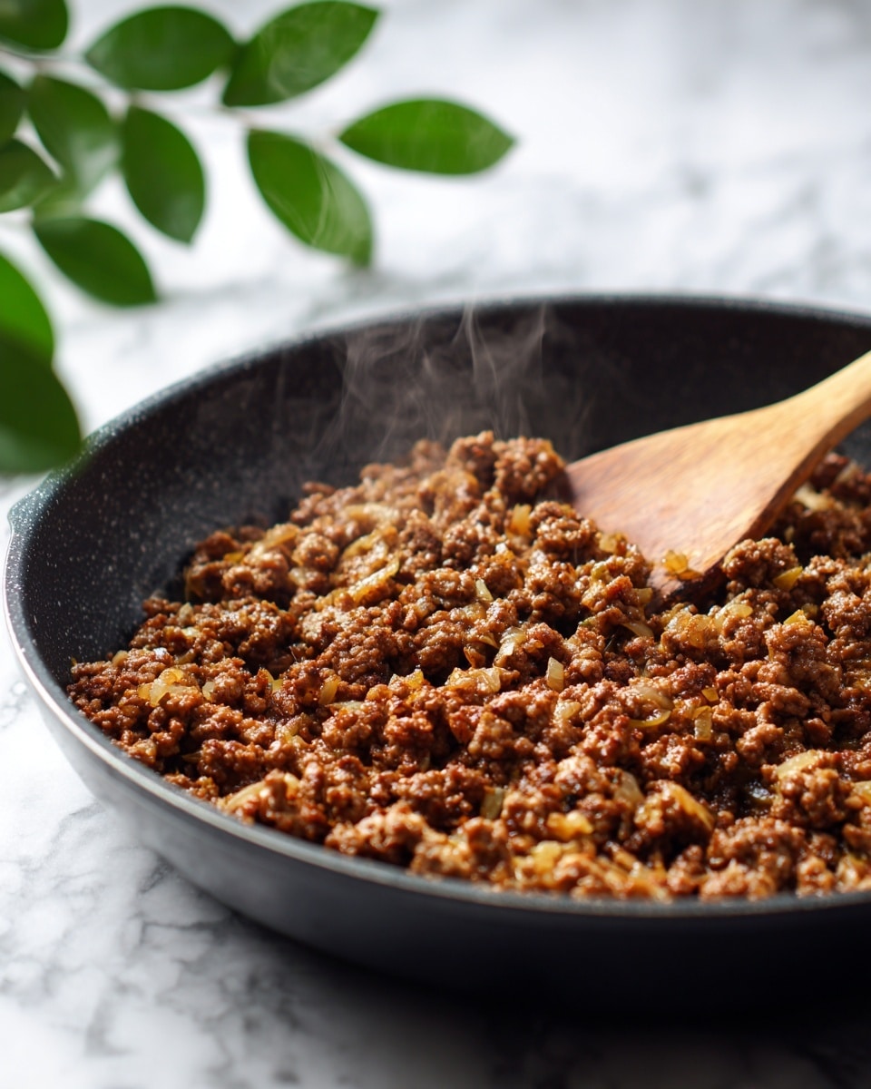 A close-up image of cooked minced meat mixed with small pieces of golden onions inside a dark skillet. The meat is richly browned with varied textures, some parts slightly crispy and others softer, showing steam rising gently. A wooden spoon rests on the right side inside the skillet, partially covered by the meat. In the blurred background, there are green leaves visible, and the whole scene is set on a white marbled surface that contrasts with the dark cooking pan. Photo taken with an iphone --ar 4:5 --v 7