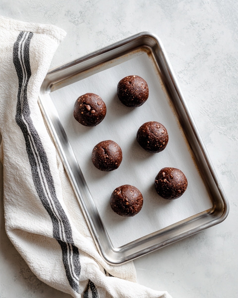 Six round dark brown cookie dough balls with visible chocolate chips are evenly spaced in two rows on white parchment paper that lines a shiny metal baking tray. The tray rests on a white marbled textured surface and a white cloth with black stripes is draped to the left side. The overall look is clean and simple with soft natural lighting. photo taken with an iphone --ar 4:5 --v 7