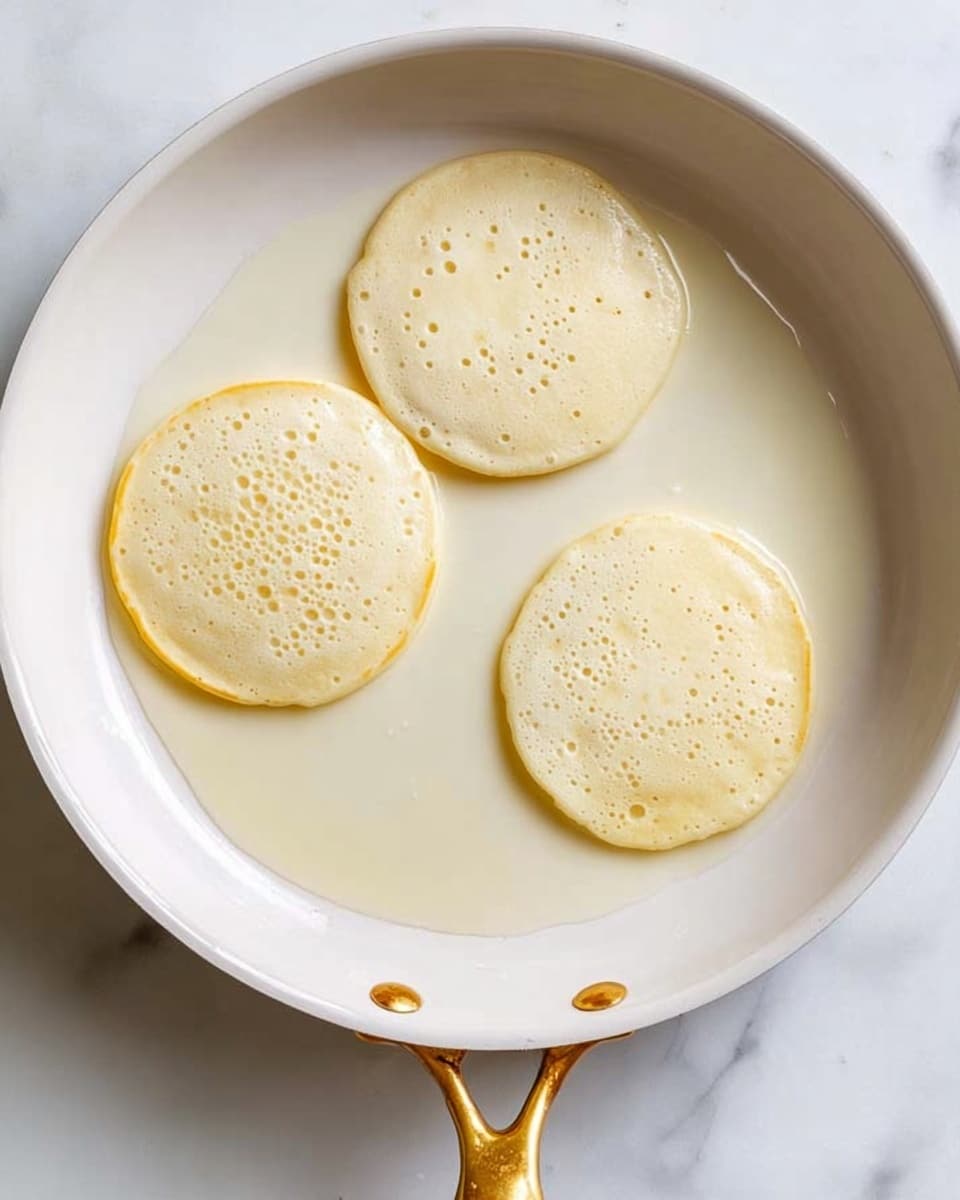 The image shows a white frying pan with a golden handle, sitting on a white marbled surface. Inside the pan are three round pancake batter pancakes cooking, each with a light yellowish color. The pancakes have small bubbles forming on the top surface and smooth, slightly shiny edges. The pan is clean and the batter looks evenly spread in uniform, circular shapes. The light highlights the smooth texture of the batter and the white pan contrasts with the golden handle. Photo taken with an iphone --ar 4:5 --v 7