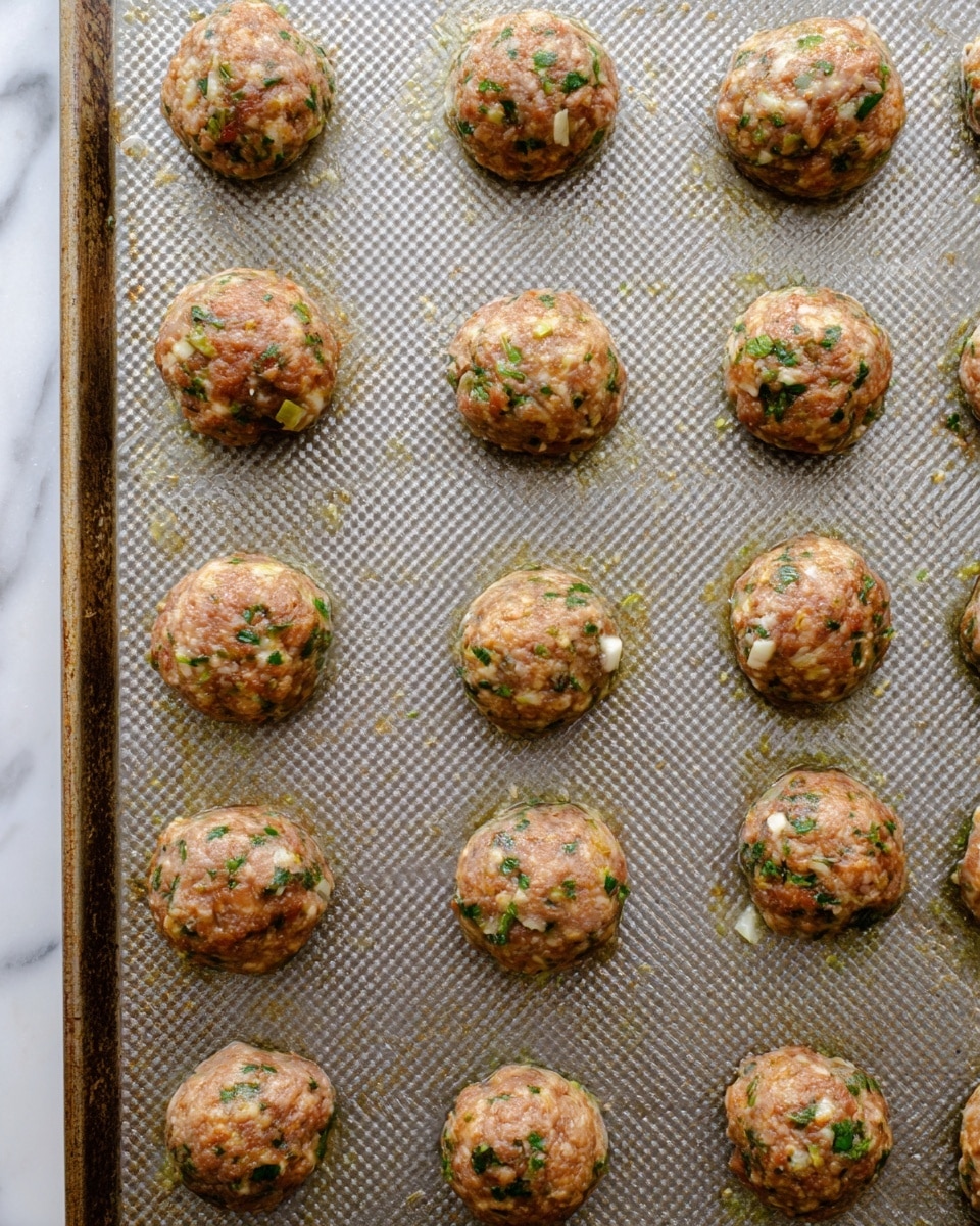 The image shows fifteen round meatballs placed evenly across a textured baking sheet with a rich grayish tone. Each meatball is light brown with visible bits of green herbs mixed throughout, alongside small white pieces that look like garlic or onion. The meatballs appear moist and slightly shiny, spaced to allow for even cooking. The baking sheet sits on a white marbled surface that contrasts with the meatballs' natural colors. photo taken with an iphone --ar 4:5 --v 7