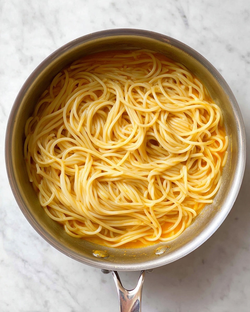The image shows a silver cooking pan filled with plain cooked spaghetti noodles on top of a smooth white marbled surface. The noodles are light yellow and are arranged in a loose swirl pattern covering a base layer of bright light orange sauce that peeks through around the edges. The pan handle is visible at the bottom, reflecting some light, and the pan's inner surface is shiny and metallic, contrasting with the soft texture of the spaghetti and sauce inside. photo taken with an iphone --ar 4:5 --v 7