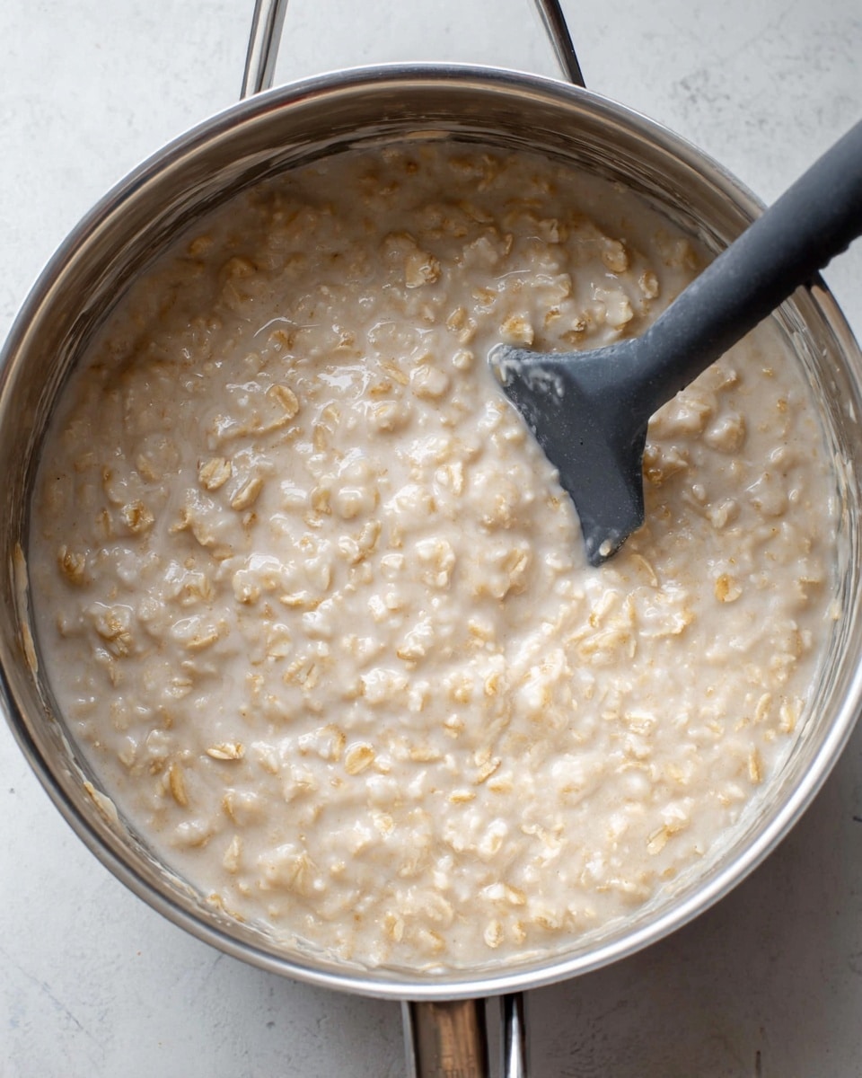 A close-up view of a cooking pot filled with thick, creamy oatmeal. The oatmeal has a light beige color with small bits of oats visible throughout, giving it a textured appearance. A black spatula with a matte finish is partially submerged on the right side, stirring the oatmeal. The pot is silver metal with a shiny finish, positioned on a white marbled surface. The oatmeal looks warm and ready to eat photo taken with an iphone --ar 4:5 --v 7
