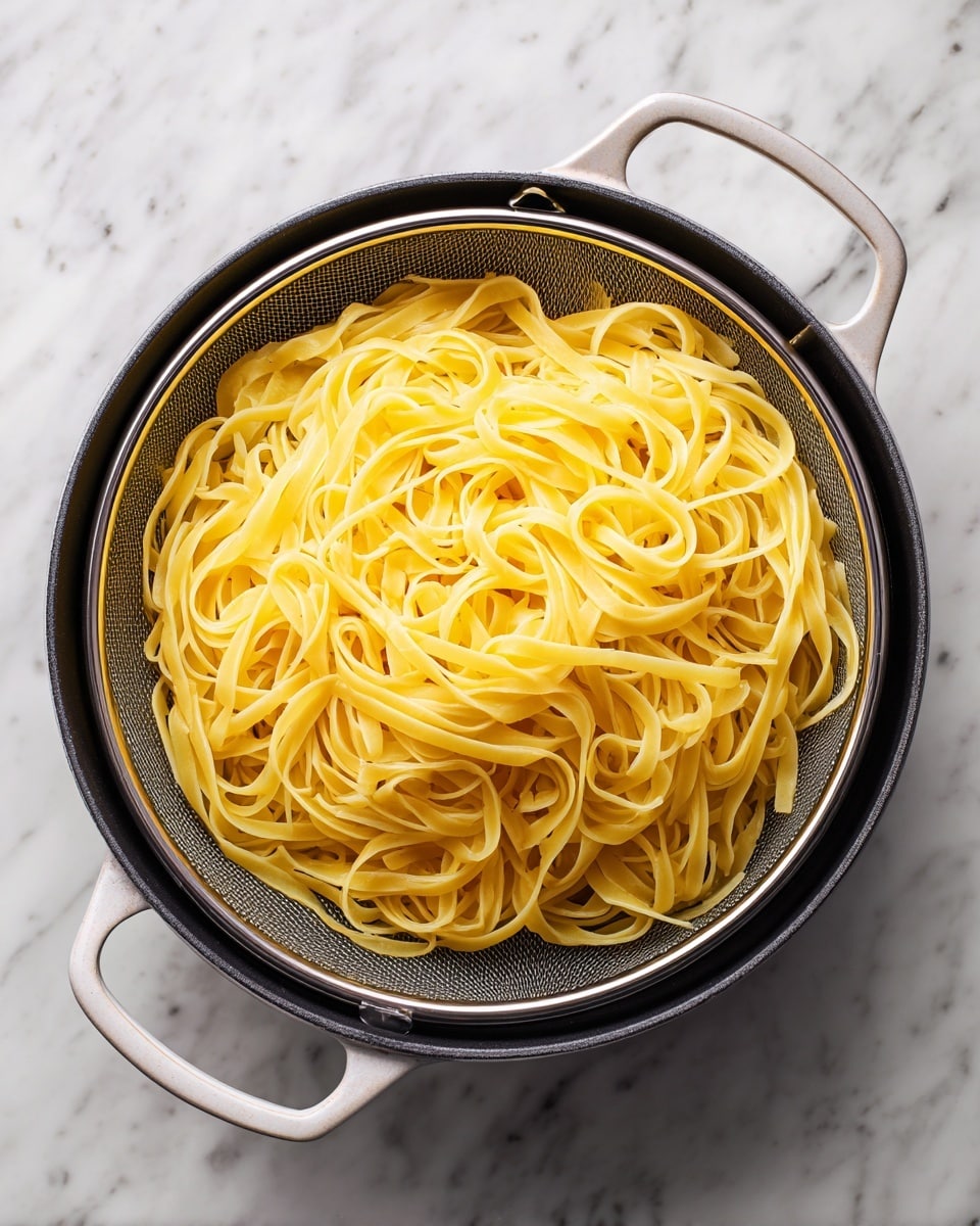 A round white pot with handles is filled with a black round pan, inside it rests a round metal sieve holding pale yellow ribbon pasta. The pasta strands are loosely piled with a smooth, slightly shiny surface and soft texture, filling the sieve completely. The background is a white marbled texture. Photo taken with an iphone --ar 4:5 --v 7