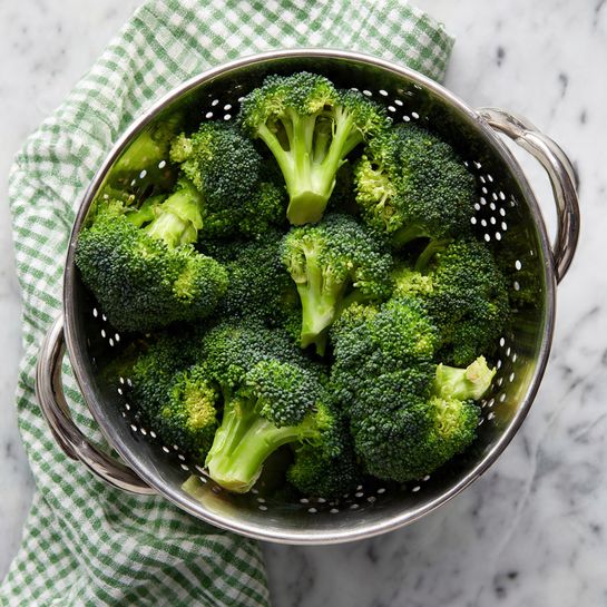 This image shows a casserole dish filled mostly with bright green broccoli pieces mixed in a creamy, pale yellow sauce that covers the broccoli well. The top layer is covered with large, spread out chunks of crispy orange crackers scattered unevenly across the surface. The edges of the casserole have a slightly browned and baked texture, showing the cheese or sauce has slightly crisped. The casserole is in a white dish, which sits on a white marbled surface. The photo taken with an iphone --ar 4:5 --v 7