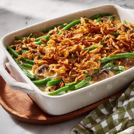 A white rectangular baking dish filled with a green bean casserole sits on a white marbled surface with a green and white checkered cloth nearby. The casserole has three visible layers: the bottom layer is bright green green beans, the middle layer shows pieces of cooked mushrooms mixed in creamy sauce, and the top layer is covered evenly with light brown crispy fried onions that add texture and crunch to the dish. The wood trivet under the dish contrasts with the clean white marbled background. Photo taken with an iphone --ar 4:5 --v 7