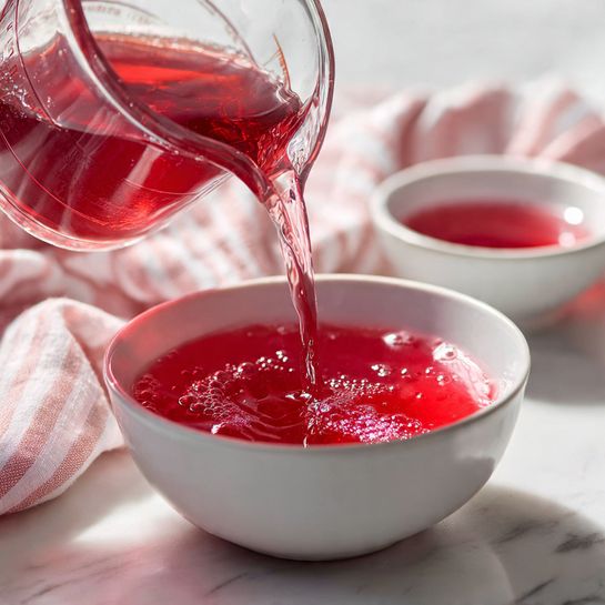 A clear glass pitcher filled with iced tea that has floating slices of strawberries near the top, showing a deep amber color with pinkish-red hues where the fruit touches the liquid. To the left, a white glass filled with the same iced tea, ice cubes, and a red-and-white striped straw, garnished with a round slice of strawberry on the rim. Around the pitcher and glass, whole strawberries and sliced strawberry pieces rest on a white marbled surface with a pink-and-white striped cloth partially visible behind the drinkware. The scene is bright and fresh with a focus on the vibrant reds and warm amber colors, photo taken with an iphone --ar 4:5 --v 7