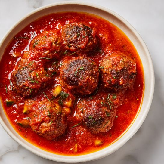 The image shows a close-up of several round, brown meatballs with a slightly rough texture, cooked with visible pieces of cooked onions embedded on the surface and sprinkled with small green herb leaves. The meatballs are placed close together inside a dark container with a shiny metal edge visible at the bottom. The lighting highlights the meatballs' moist and textured surface, and the background shows a white marbled texture. photo taken with an iphone --ar 4:5 --v 7
