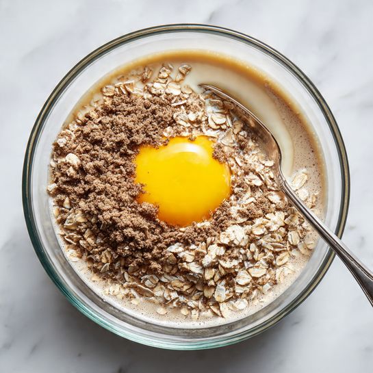 A clear glass bowl on a white marbled surface holds a mixture of three layers. The bottom layer is light brown liquid with a foamy texture. The middle layer is a large amount of dry oats mixed with brown powder, giving a rough texture and dark beige color. The top layer is a bright yellow, slightly mixed egg yolk sitting in the center of the oats. A silver spoon is partially visible in the bowl, placed on the right side, with its edge touching the oats and liquid. Photo taken with an iphone --ar 4:5 --v 7