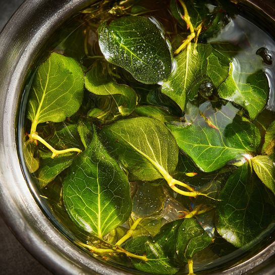 A tall clear glass filled with light golden iced tea sits in front, containing many small ice pieces with two fresh green mint leaves on top and a pink and white striped straw inside. Behind it, a clear glass pitcher holds more iced tea with floating lemon slices and mint leaves, positioned on a worn wooden table with a folded blue and white striped cloth to the left and a few loose green mint leaves scattered nearby. The background is a white tiled wall, and the surface beneath everything has a white marbled texture. photo taken with an iphone --ar 4:5 --v 7