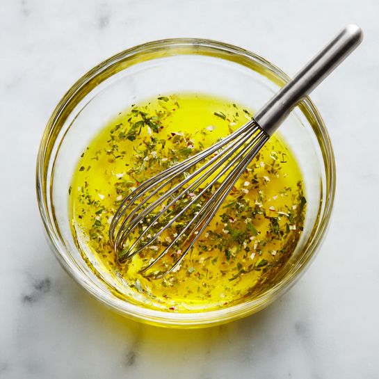 A clear glass bowl holds a bright yellow oil-based mixture with small herb flakes floating inside. A silver metal whisk rests in the bowl, partially submerged in the liquid. The bowl sits on a white marbled surface, creating a clean and simple background. The photo has soft, natural lighting that highlights the smooth texture of the oil and the metallic shine of the whisk. photo taken with an iphone --ar 4:5 --v 7