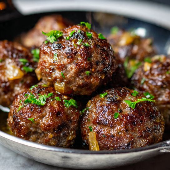 The image shows a close-up of a rich, thick tomato sauce with multiple browned meatballs partially submerged in it. The sauce is bright red with visible chunks of cooked onions, green and yellow zucchini pieces, and small bits of herbs, giving it a textured look. The meatballs have a browned, slightly rough surface with some sauce coating them, and the sauce appears glossy with some small pools of oil reflecting light. The background is a white marbled texture photo taken with an iphone --ar 4:5 --v 7