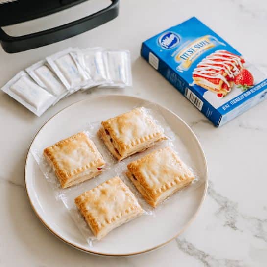 The image shows a white plate on a white marbled surface with a clear plastic package containing four light golden toaster strudel pastries, each with a flaky and slightly crispy texture. Above the pastries, there are six small rectangular white frosting packets lined up neatly. To the top right of the plate, there is a blue box of Pillsbury Toaster Strudel strawberry pastries featuring the Pillsbury Doughboy and a picture of a strudel with white icing drizzle and red strawberry filling. Part of a black air fryer basket with a handle is visible on the top left corner. The overall scene is bright and clean with soft natural lighting photo taken with an iphone --ar 4:5 --v 7