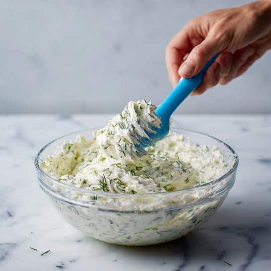 The image shows a clear glass bowl filled with a creamy white mixture that has small green herb pieces mixed throughout, giving it a textured look. A blue silicone spatula is partially in the mixture, lifting and folding it, while a woman's hand is visible holding the bowl from the side. The background and surface beneath the bowl have a white marbled texture. The creamy mixture appears soft and fluffy, with visible bits of green herbs spread evenly inside. Photo taken with an iphone --ar 4:5 --v 7