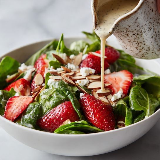 A close-up view of a fresh salad in a white bowl placed on a white marbled surface, showing three main layers: a base of bright green leafy spinach and arugula leaves with smooth and slightly ruffled textures, scattered thinly sliced red strawberries with juicy, glossy surfaces on top, and toasted thin almond slices with light brown crisp edges sprinkled over the strawberries. Small white crumbles of soft cheese are spread lightly across the salad. A woman's hand is pouring a creamy light beige dressing speckled with black pepper over the salad from a speckled ceramic pitcher. The image focuses on the vibrant colors and textures of the fresh ingredients and flowing dressing photo taken with an iphone --ar 4:5 --v 7