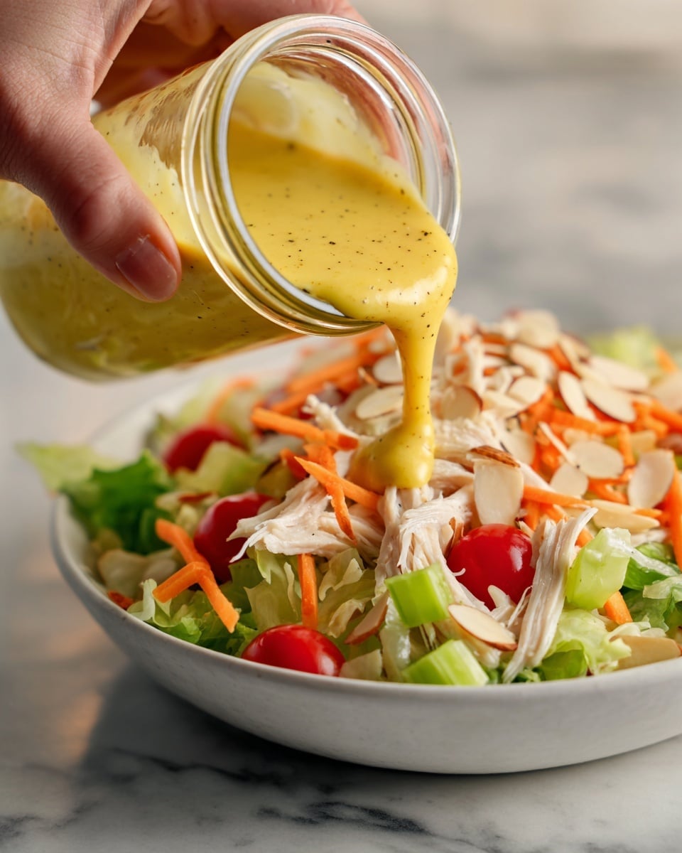A close-up image shows a woman's hand pouring a thick yellow dressing with black specks from a clear glass jar onto a colorful salad. The salad beneath has several visible layers, including shredded white pieces of chicken on top, bright green chopped celery, thin orange carrot shreds, slivered white almonds, and small red tomato pieces mixed in. The salad sits in a white bowl on a white marbled surface, with the background softly blurred to keep focus on the jar and salad. Photo taken with an iphone --ar 4:5 --v 7