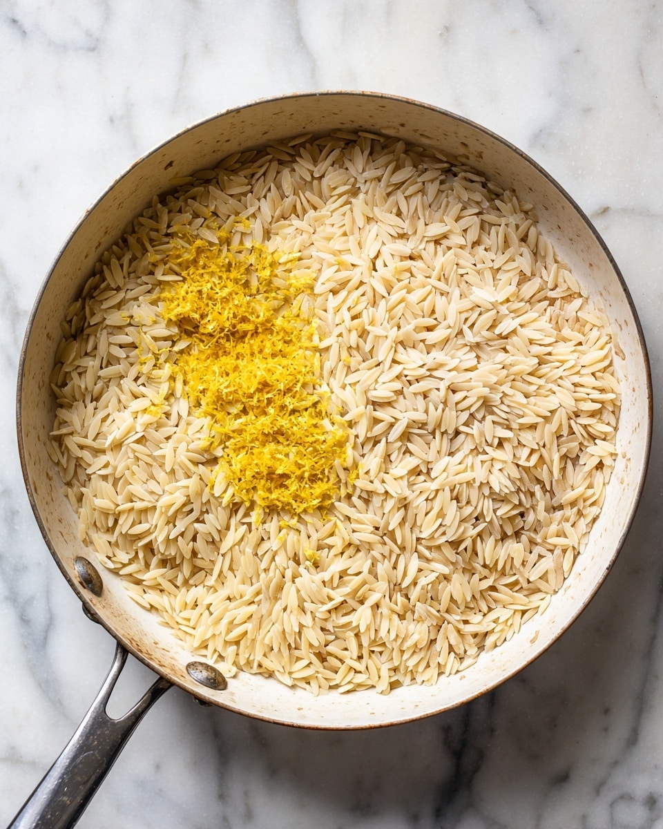 A large white pan viewed from above holds a single layer of uncooked orzo pasta. The orzo grains are spread evenly, showing a light golden brown color with some parts slightly toasted. On one side of the pan, there is a small pile of finely grated yellow lemon zest resting gently on top of the orzo. The pan is placed on a white marbled surface, creating a clean and bright setting. photo taken with an iphone --ar 4:5 --v 7