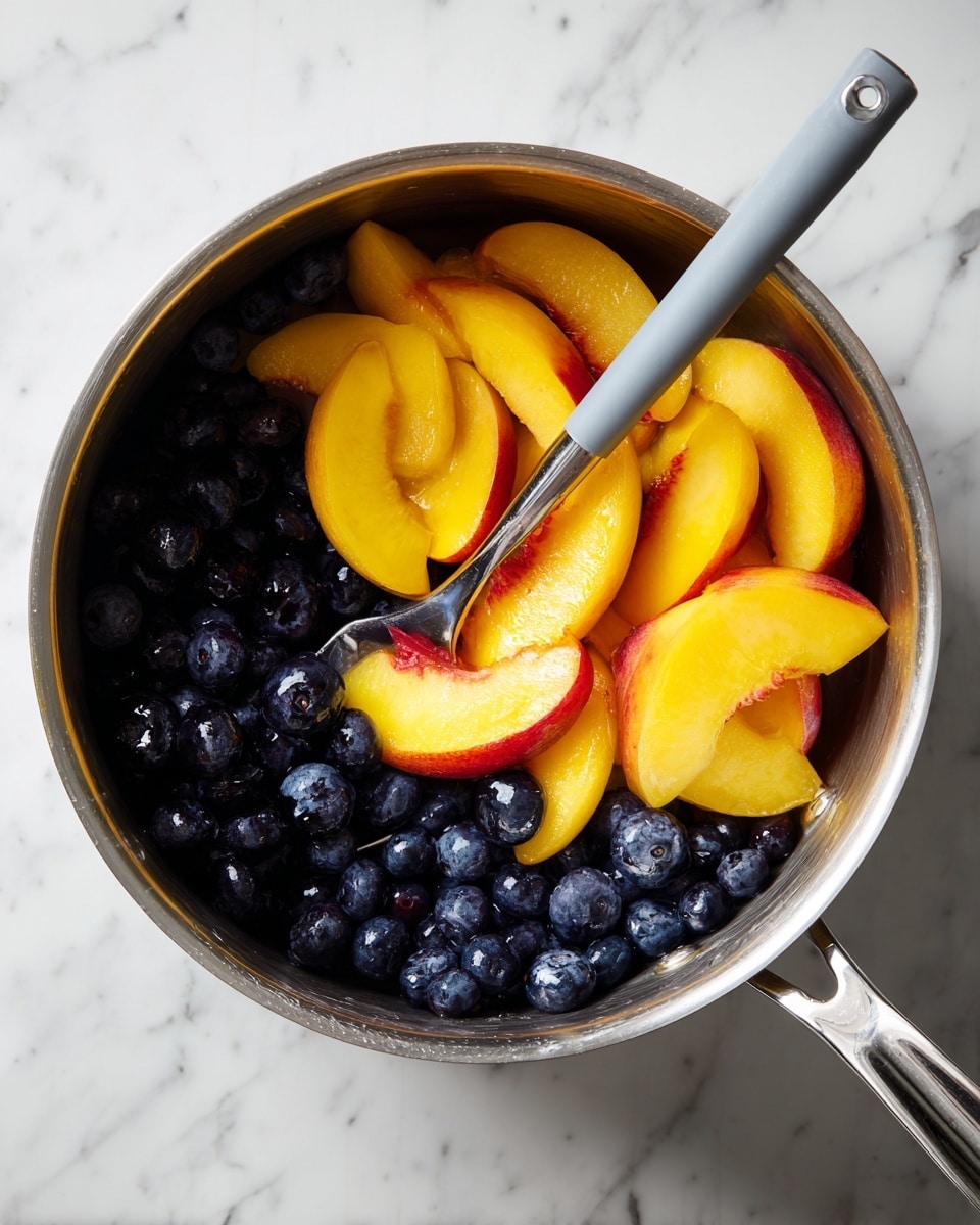 A metal pot filled with fresh fruit slices is shown from above. The bottom layer of the pot contains small, round dark blueberries, and the upper layer consists of several thick yellow-orange peach slices with some red highlights on the edges. A silver-handled spatula with a gray silicone head is partially submerged among the fruit, resting inside the pot. The pot is placed on a surface with a white marbled texture. photo taken with an iphone --ar 4:5 --v 7