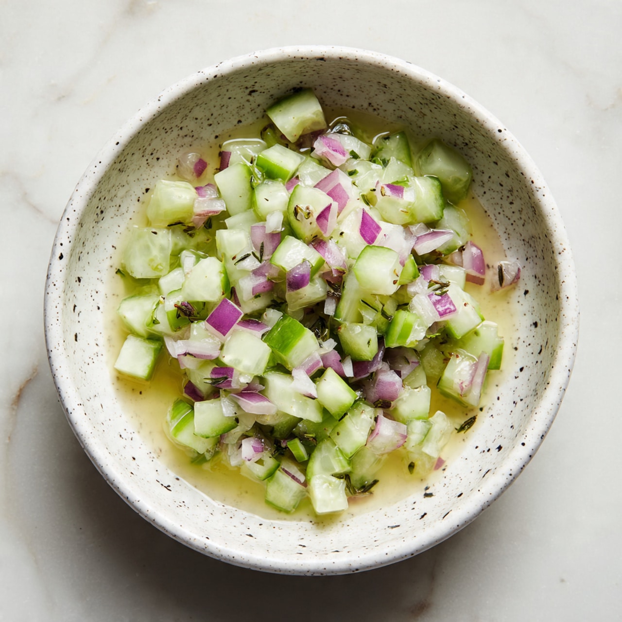 A shallow white speckled bowl holds finely diced light green cucumbers and light purple shallots layered evenly, floating in a clear, slightly yellow liquid that appears as light dressing. The small cubes of vegetables are evenly spread across the bowl, showing contrast between the green and purple hues against the creamy white bowl with black speckles. The bowl sits on a white marbled textured surface, creating a clean and fresh look. Photo taken with an iphone --ar 4:5 --v 7