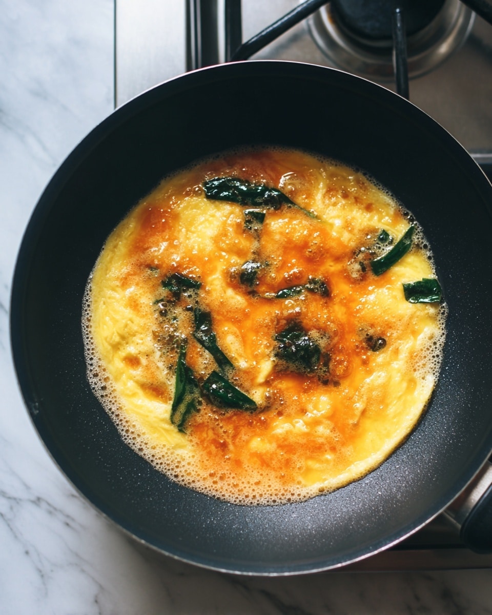 The image shows an egg omelet cooking in a black pan, with a thin yellow layer of cooked egg forming the base. Over the egg, there is a spread of orange sauce with a slightly shiny and smooth texture, bubbling gently. There are scattered dark green leafy vegetables, some partially submerged in the sauce and some resting on top of the egg layer. The pan sits on a stove with metallic elements visible in the background, and the surface beneath the stove has a white marbled texture. photo taken with an iphone --ar 4:5 --v 7