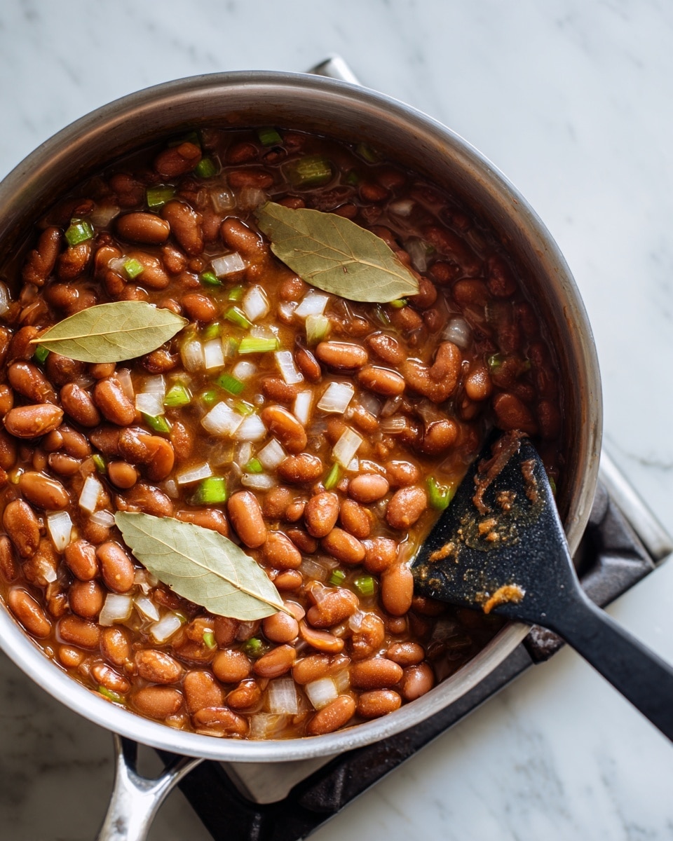 A close-up view of a stainless steel pot filled with cooked reddish-brown beans mixed with small diced translucent white onions and green bell pepper pieces. Two light beige bay leaves lay on top, adding texture and color contrast. A black spatula with some food stains rests on the right side inside the pot. The pot sits on a gas stove burner against a white marbled surface. Photo taken with an iphone --ar 4:5 --v 7
